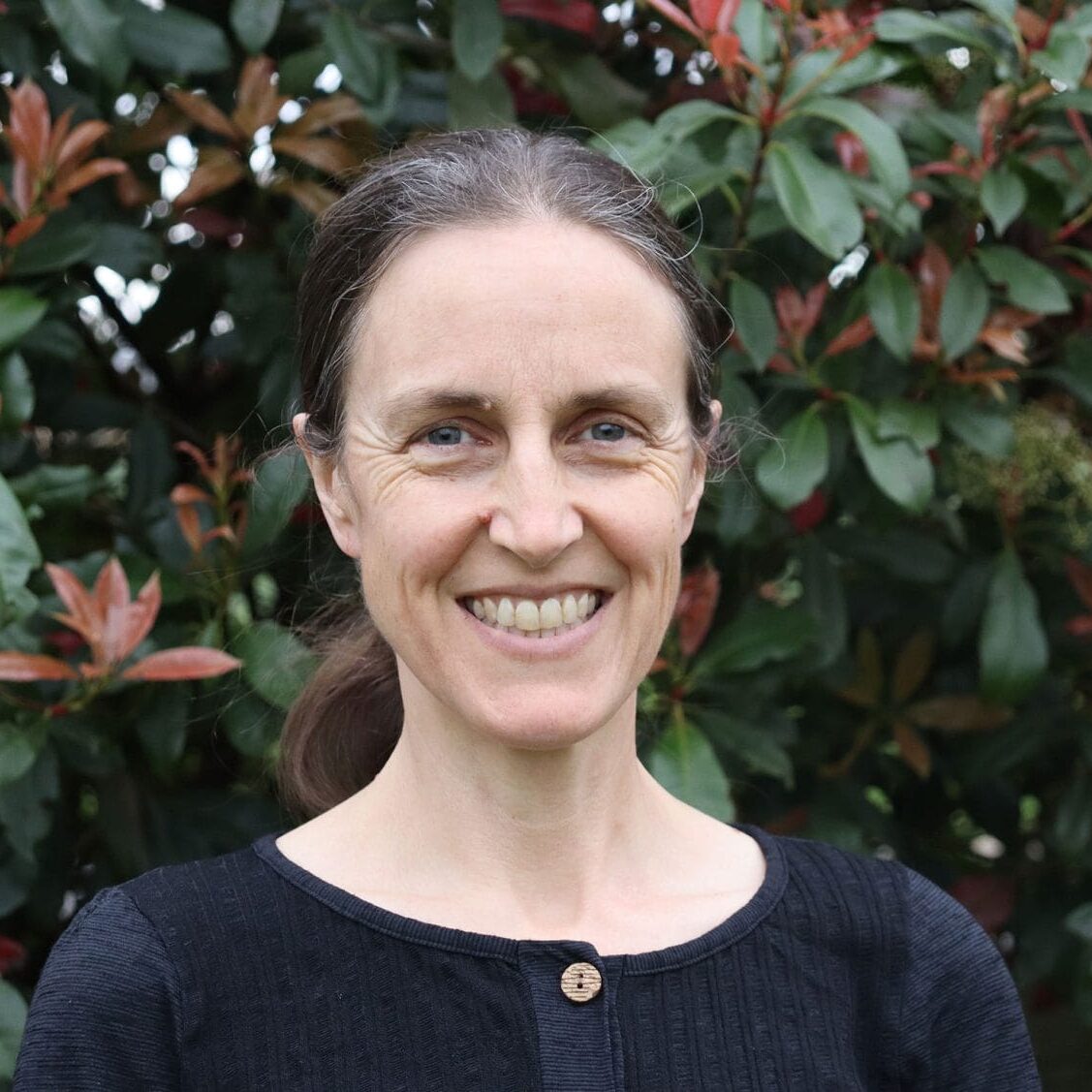 Smiling woman with dark hair in a dark top, standing in front of green and red-tinged shrubbery outdoors.
