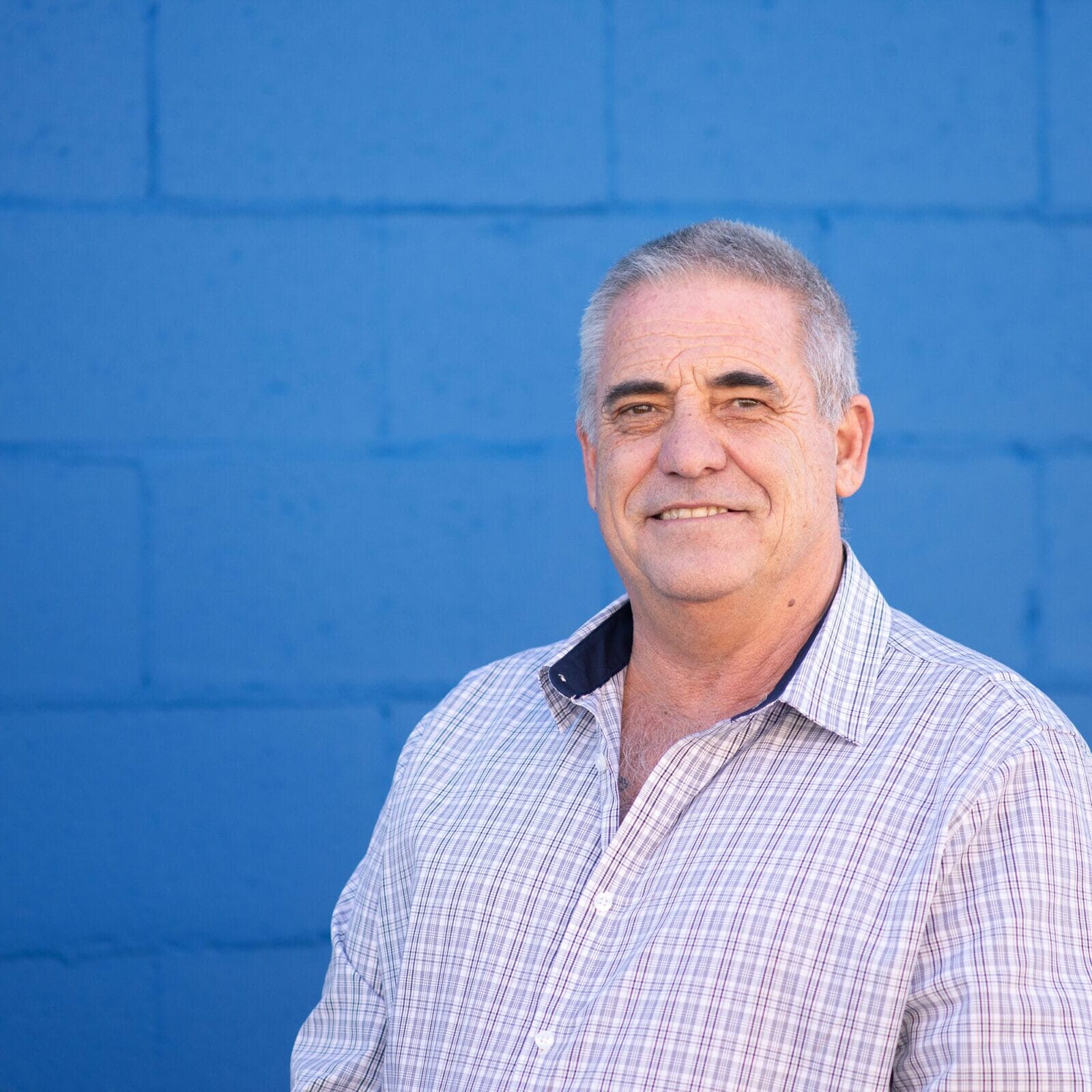 Smiling older man with short gray hair wearing a light plaid shirt, standing in front of a blue wall.