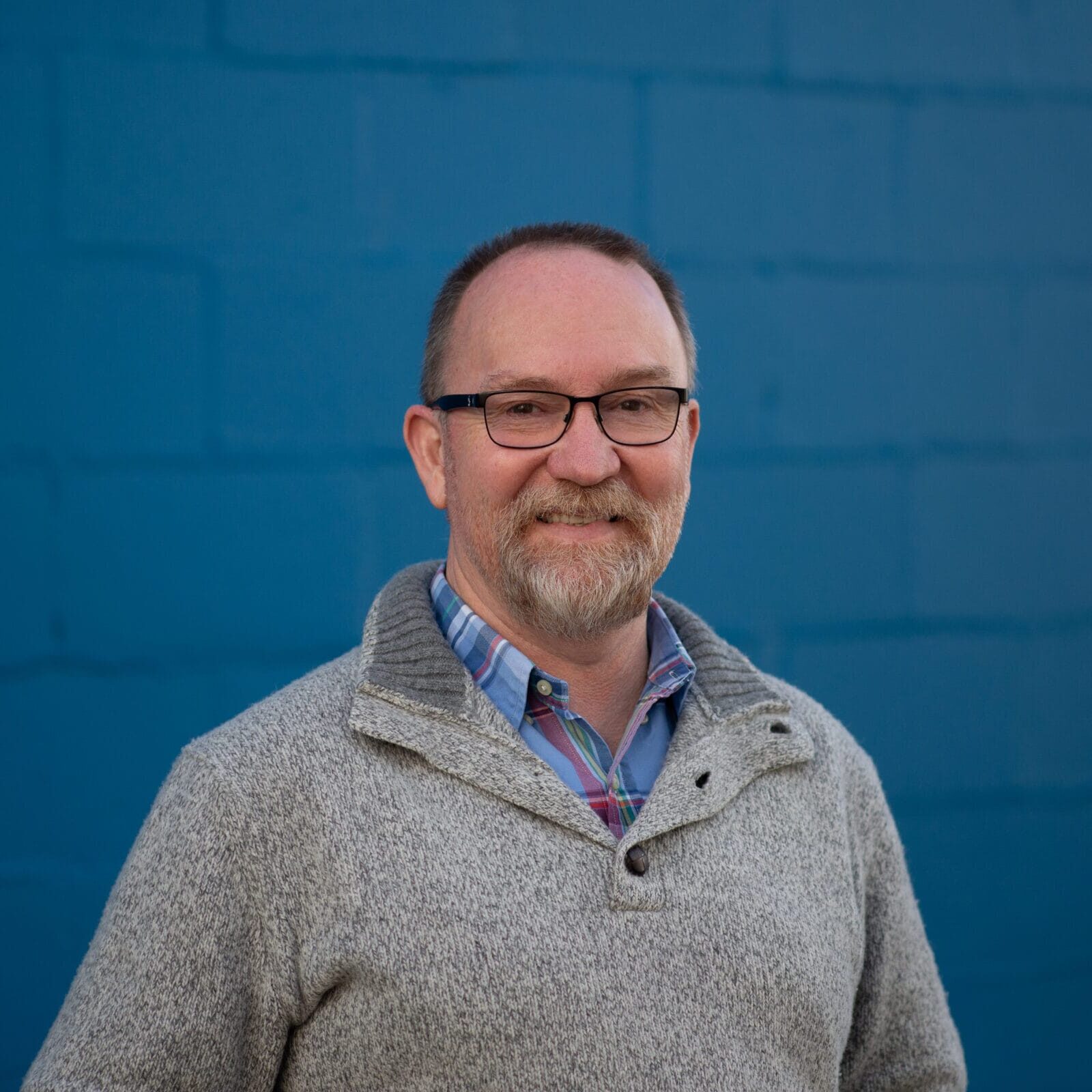Smiling man with glasses and a gray beard, wearing a plaid shirt and gray sweater, posed against a blue wall.