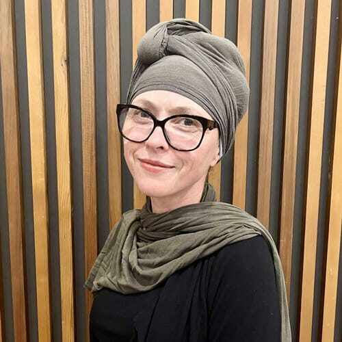 Portrait of a woman wearing a gray turban and olive scarf, smiling at the camera against a wooden striped background with black-frame glasses.